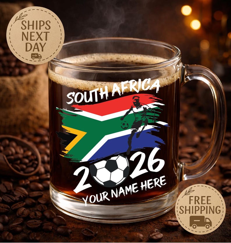 May include: A clear glass coffee mug filled with dark coffee. The mug features a design with the South African flag, a soccer player, a soccer ball, and the text "SOUTH AFRICA 2026 YOUR NAME HERE".