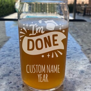 May include: A clear glass with a graduation cap design and the text "I'm Done" with a banner underneath. The glass is filled with a light brown liquid and has the text "Custom Name Year" at the bottom.