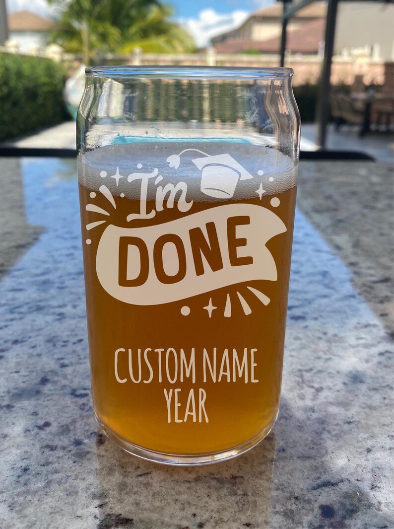 May include: A clear glass with a graduation cap design and the text "I'm Done" with a banner underneath. The glass is filled with a light brown liquid and has the text "Custom Name Year" at the bottom.