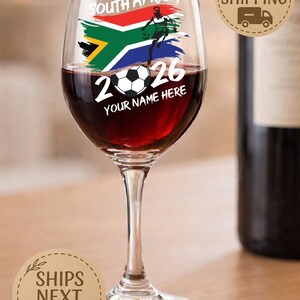 May include: A wine glass filled with red wine, featuring a design with the South African flag, a soccer player, and the text "2026 YOUR NAME HERE". The glass is on a wooden surface.