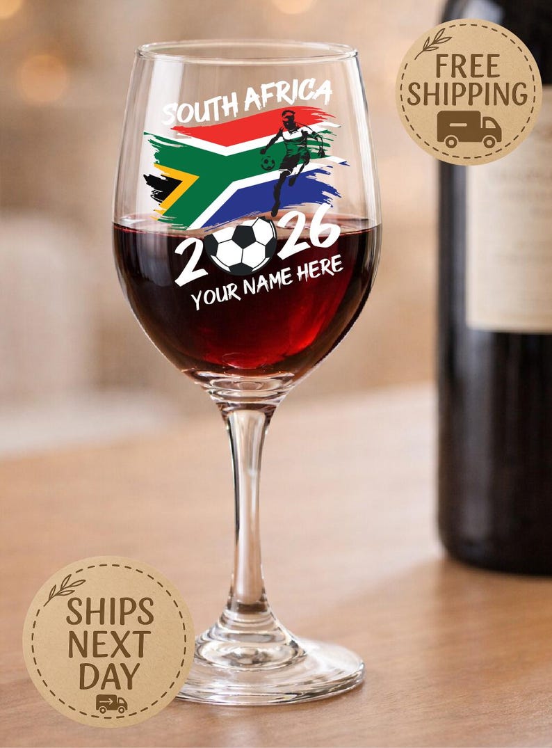 May include: A wine glass filled with red wine, featuring a design with the South African flag, a soccer player, and the text "2026 YOUR NAME HERE". The glass is on a wooden surface.
