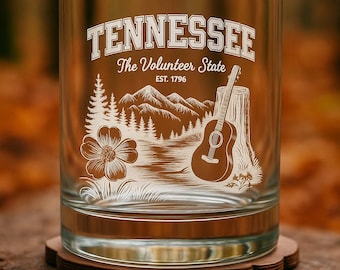 May include: A clear glass tumbler with a Tennessee-themed design. The design includes the word "Tennessee", the phrase "The Volunteer State", a mountain scene, a guitar, and a flower. The glass sits on a wooden coaster.