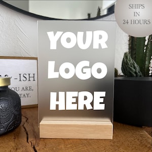 May include: A clear acrylic sign with the words "YOUR LOGO HERE" in white, set on a natural wood base. The sign is displayed on a wooden surface, with a decorative bottle and a small sign in the background.