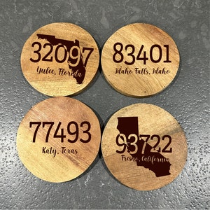 May include: Four round wooden coasters with engraved text and state outlines. Each coaster features a different zip code and city: 32097 Yulee, Florida; 83401 Idaho Falls, Idaho; 77493 Katy, Texas; and 93722 Fresno, California.