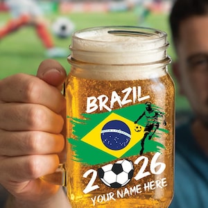 May include: A glass beer mug filled with beer and foam, decorated with a Brazilian flag design and the word "BRAZIL". It also features a soccer player graphic, a soccer ball, and the numbers "2026". The words "YOUR NAME HERE" are also visible on the mug.