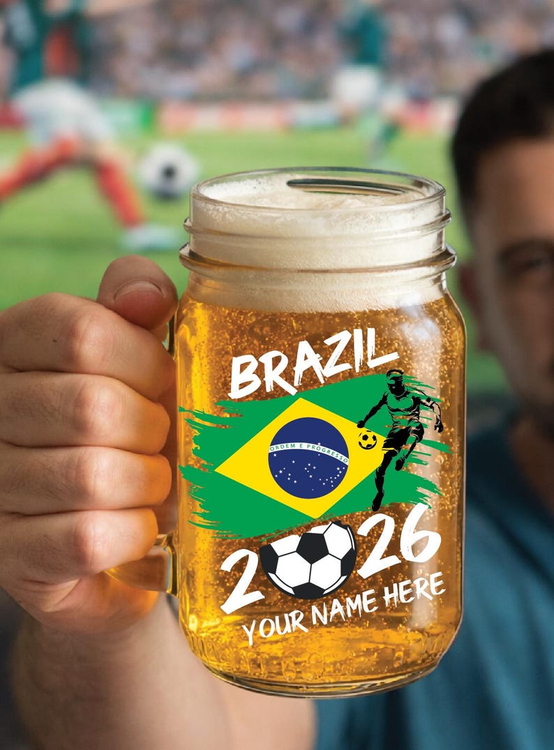 May include: A glass beer mug filled with beer and foam, decorated with a Brazilian flag design and the word "BRAZIL". It also features a soccer player graphic, a soccer ball, and the numbers "2026". The words "YOUR NAME HERE" are also visible on the mug.