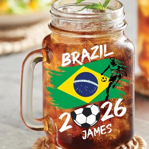 May include: A glass jar mug filled with a brown beverage, ice, and a sprig of mint. The mug features the word "BRAZIL" and a Brazilian flag graphic. Below the flag are the numbers "2026", a soccer ball, and the name "JAMES".