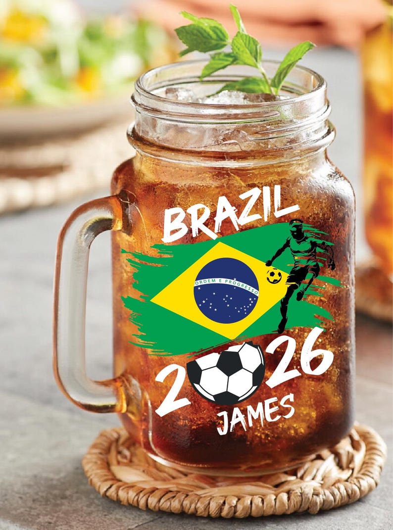 May include: A glass jar mug filled with a brown beverage, ice, and a sprig of mint. The mug features the word "BRAZIL" and a Brazilian flag graphic. Below the flag are the numbers "2026", a soccer ball, and the name "JAMES".
