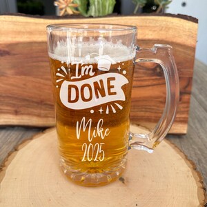 May include: A clear glass beer mug with a white design that says "I'm Done" with a graduation cap, fireworks, and a banner. The mug also says "Mike 2025".