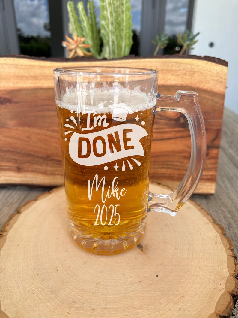 May include: A clear glass beer mug with a white design that says "I'm Done" with a graduation cap, fireworks, and a banner. The mug also says "Mike 2025".