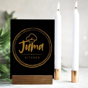 May include: A black sign with the gold text "Juma Kitchen" and a chef's hat logo, held in a wooden stand. Two white candles with gold bases are lit, creating a warm ambiance. The scene suggests a restaurant or kitchen setting.