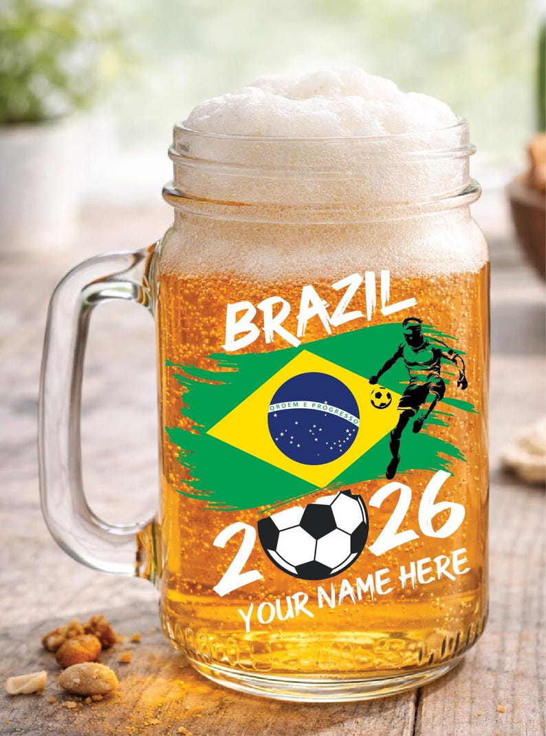 May include: A clear glass beer mug filled with beer and foam. The mug features a Brazilian flag design, a soccer player graphic, and the text "BRAZIL 2026". The bottom text reads "YOUR NAME HERE".