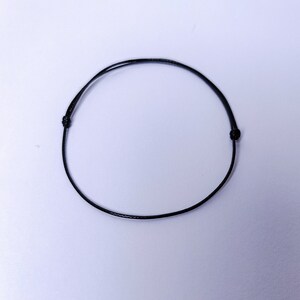 Puede incluir: Una pulsera negra minimalista hecha de un cordón fino y flexible. La pulsera tiene nudos en ambos extremos para ajustar el tamaño. El cordón es de color negro intenso y la pulsera se muestra sobre un fondo blanco.