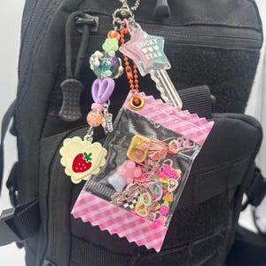 May include: A keychain featuring a clear plastic pouch filled with colorful miniature charms, a star-shaped charm with the text LUCKY STAR, a strawberry-shaped charm, and a strand of multi-colored beads attached to a metal ring.