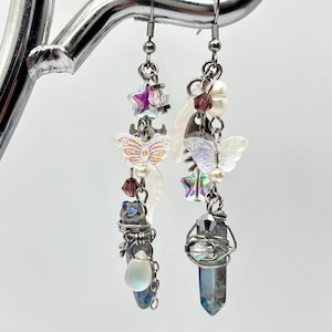 May include: A pair of long, dangling earrings with a whimsical, ethereal theme. Each earring features a blue iridescent crystal point, a white iridescent butterfly, a crescent moon charm, a star charm, and a variety of iridescent beads.