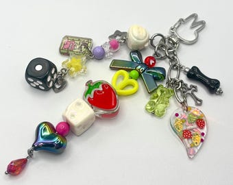 May include: A collection of charms on a silver chain, including a black die, a red strawberry, a cross, a bone, and a heart-shaped charm with iridescent colors. Other charms include a yellow star, a gummy bear, and a tooth.
