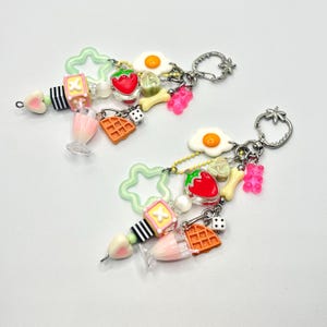 May include: A pair of dangle earrings with a whimsical breakfast theme. Each earring features a variety of charms, including a fried egg, a strawberry, a waffle, a milkshake, a star, a bone, a dice, and a gummy bear. The charms are attached to a silver chain and a silver hook.