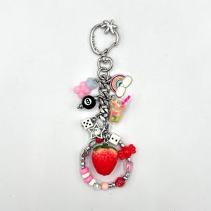 May include: A silver charm keychain with a variety of colorful charms, including a rainbow, a strawberry, a dice, a cloud, a pink and blue donut, a black 8 ball, and a pink and white drink with a straw.