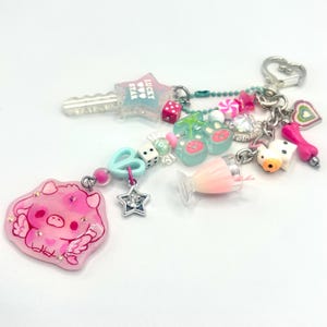 May include: A whimsical keychain with a variety of charms. It features a pink pig charm, a star-shaped key, dice, cherries, a milkshake, and other decorative elements. The keychain is attached to a silver-colored clasp.