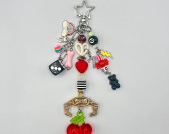 May include: A silver keychain with a star-shaped clasp, adorned with various charms. Charms include a rainbow, dice, a pink bone, a pretzel, a red heart, a black and white striped bead, a gold claw machine with the word "YOU", and red cherries.