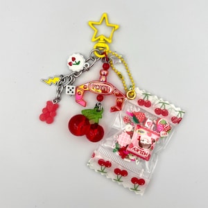 May include: A yellow star-shaped keychain featuring a red claw machine charm with the text YOUYI, a red gummy bear, a white die, a lightning bolt, a cherry charm, and a clear plastic bag filled with small fruit-themed charms and a Milky candy packet.