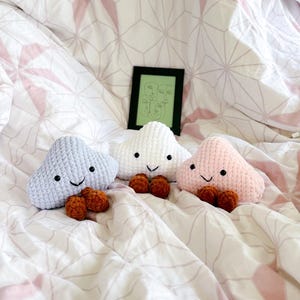 Könnte beinhalten: Drei gehäkelte Wolken-Plüschtiere in Grau, Weiß und Rosa mit braunen Knopfaugen und braunen Garnnasen. Die Spielzeuge liegen auf einer weißen und rosa gemusterten Bettdecke.