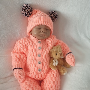 May include: A pink knitted baby onesie with a matching pink and black pom-pom hat. The onesie has a button closure and a teddy bear is lying next to the baby.