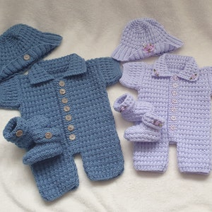 Può includere: Due completi per neonati lavorati a maglia, blu e lavanda. Ogni set include una tutina con bottoni in legno, un cappello e scarpine abbinate. L'outfit lavanda ha piccoli accenti floreali. I completi sono esposti su una superficie bianca.