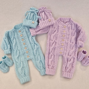 May include: Two knitted baby outfits in light blue and lilac. Each set includes a romper with wooden buttons, a matching hat, and booties. The rompers feature a cable knit design.