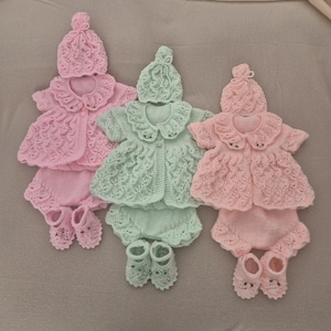 Puede incluir: Tres conjuntos de ropa de bebé de punto en tonos pastel. Cada conjunto incluye un gorro, una chaqueta de manga corta con cuello festoneado, un pantalón corto y patucos. Los conjuntos son de color rosa y verde menta, todos tejidos a mano con un patrón texturizado.