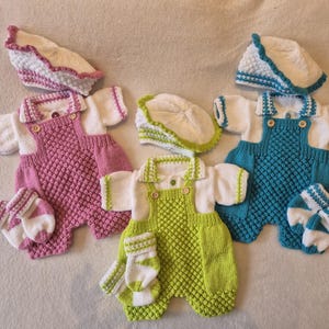 May include: Three knitted baby outfits in pink, green, and teal. Each outfit includes a romper with white sleeves, a matching hat, and mittens. The rompers have a textured stitch and wooden buttons.