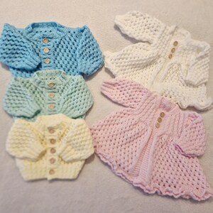 May include: Five handmade baby cardigans in pastel colors: light blue, white, pink, yellow, and mint green. Each cardigan features a button-up front and a textured crochet design. The cardigans are arranged on a soft, neutral surface.