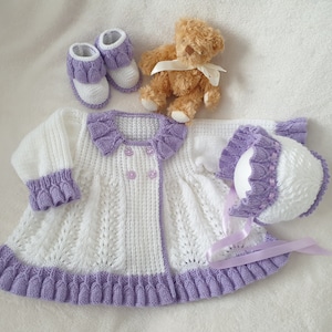 May include: A white and lavender knitted baby outfit set, including a coat, bonnet, and booties. A small teddy bear with a cream ribbon is also present. The set is made of soft yarn and features a detailed pattern.