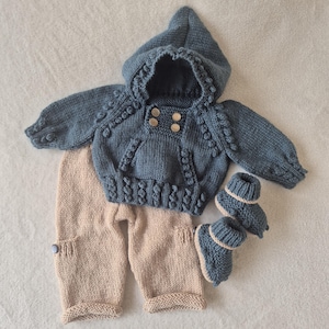 May include: A knitted baby outfit in blue and beige. The set includes a hooded sweater with wooden buttons, trousers, and matching booties. The sweater has decorative bobbles and the trousers have a button detail on the pocket.