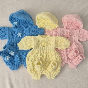 May include: Three knitted baby outfits in blue, yellow, and pink. Each outfit includes a matching hat and booties. The outfits are made with a delicate cable knit pattern and feature flower embellishments.