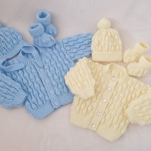 Peut inclure: Deux ensembles tricotés pour bébé, l'un en bleu clair et l'autre en crème. Chaque ensemble comprend un cardigan, un bonnet et des chaussons.