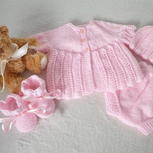 May include: A pink knitted baby outfit set including a cardigan, pants, and booties. The set is made with a delicate stitch pattern and features a button closure on the cardigan.