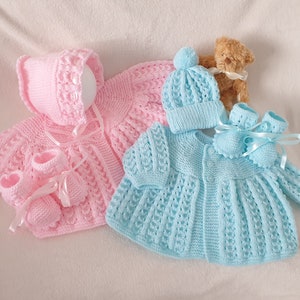 May include: Two knitted baby outfits, one pink and one blue, with matching hats and booties. The outfits are made with a delicate lace pattern and feature button closures.