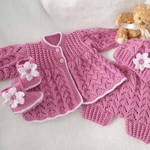 Puede incluir: Un conjunto de bebé de punto rosa con un cárdigan, pantalones, gorro y botines. El conjunto tiene un ribete blanco y detalles de flores blancas.