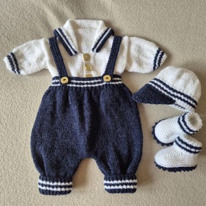May include: A knitted baby outfit consisting of a white shirt with a sailor collar, navy blue dungarees with suspenders, a matching white and navy cap, and two white booties. The shirt features three gold buttons and navy striped trim on the sleeves.