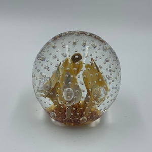 May include: A clear glass paperweight, spherical in form. It contains amber-coloured, petal-like designs within. Numerous small air bubbles are visible throughout the glass. The base of the paperweight has a darker amber tone. A decorative item.