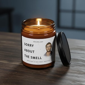 May include: A lit amber glass candle jar with a black lid. The label reads "SORRY ABOUT THE SMELL" with a picture of a man. The candle is made of 100% natural soy wax.