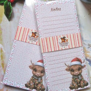 Könnte beinhalten: Zwei rechteckige Notizblöcke mit einem Rand aus Zuckerstangenmustern und einer Grafik eines Hochlandrindes mit Weihnachtsmütze und Lichterkette. Ein Papierband mit dem Text Wickingly Delicious und einer Katzenillustration umschließt jeden Block.