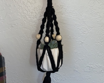 May include: Black macrame plant hanger with wooden beads and a white ceramic pot. The hanger is hanging from a white wall.