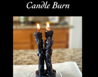 May include: Two black candles in the shape of a couple standing together, with flames burning at the top. The candles are on a glass plate with a heart-shaped base. Two pink crystals are on a white surface below the candles.