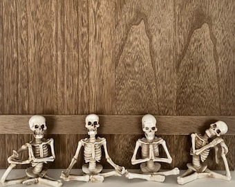 May include: Four white skeleton figurines in a seated yoga pose. The skeletons are arranged in a row on a white surface with a brown wooden background.