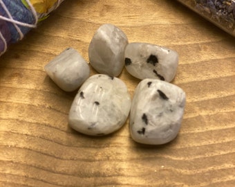 May include: Five white moonstone tumbled stones with black inclusions. The stones are on a wooden surface.
