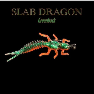 May include: A green and orange Slab Dragon Greenback fishing lure. The lure has a segmented body, multiple legs, and a hook. The text "SLAB DRAGON" and "Greenback" are displayed above the lure on a black background.