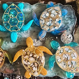 Pode incluir: Uma coleção de tartarugas decorativas com conchas preenchidas com pequenas pedras. As tartarugas são de várias cores, incluindo verde, azul e dourado. Também existem cruzes decorativas com as palavras "Faith" e "Blessed".