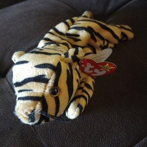 Op de afbeelding: Een pluche tijger beanie baby speelgoed met zwarte en witte strepen. Het speelgoed heeft een rood en wit label met de tekst "Beanie Baby" met een ster.